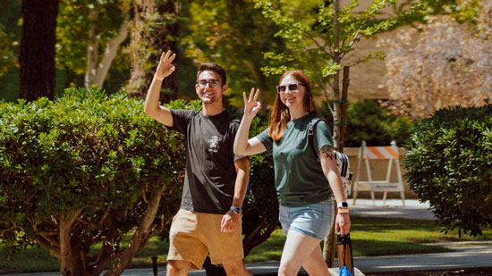 Two students walking in front of bushes and showing the Rowdy symbol with their hands